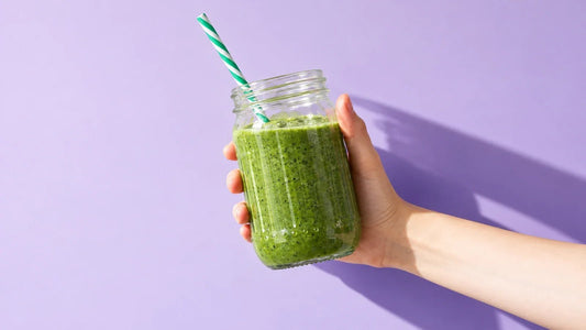 Hand holding a green Spirulina smoothie against a sunlit lavender background.